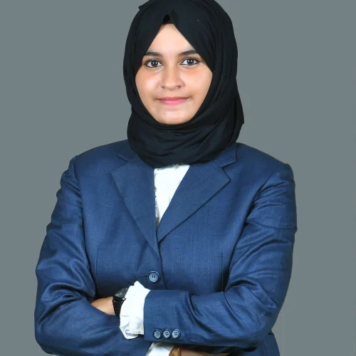 Professional woman wearing a black hijab and blue blazer with arms crossed against a plain background Caption: